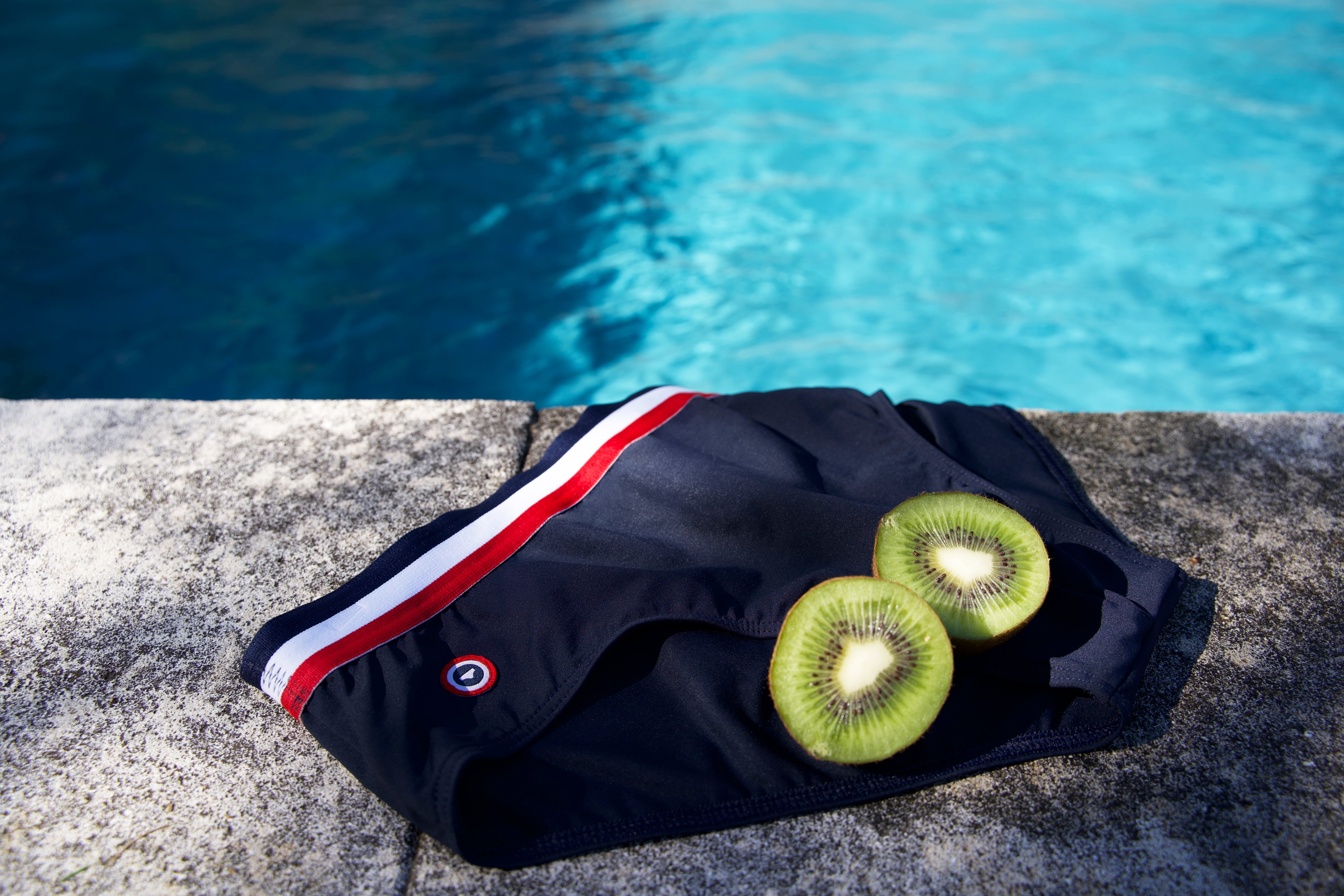 Slip de bain bleu marine avec ceinture rayure tricolore rouge et blanche posé au bord d'une piscine avec deux tranches de kiwi frais.