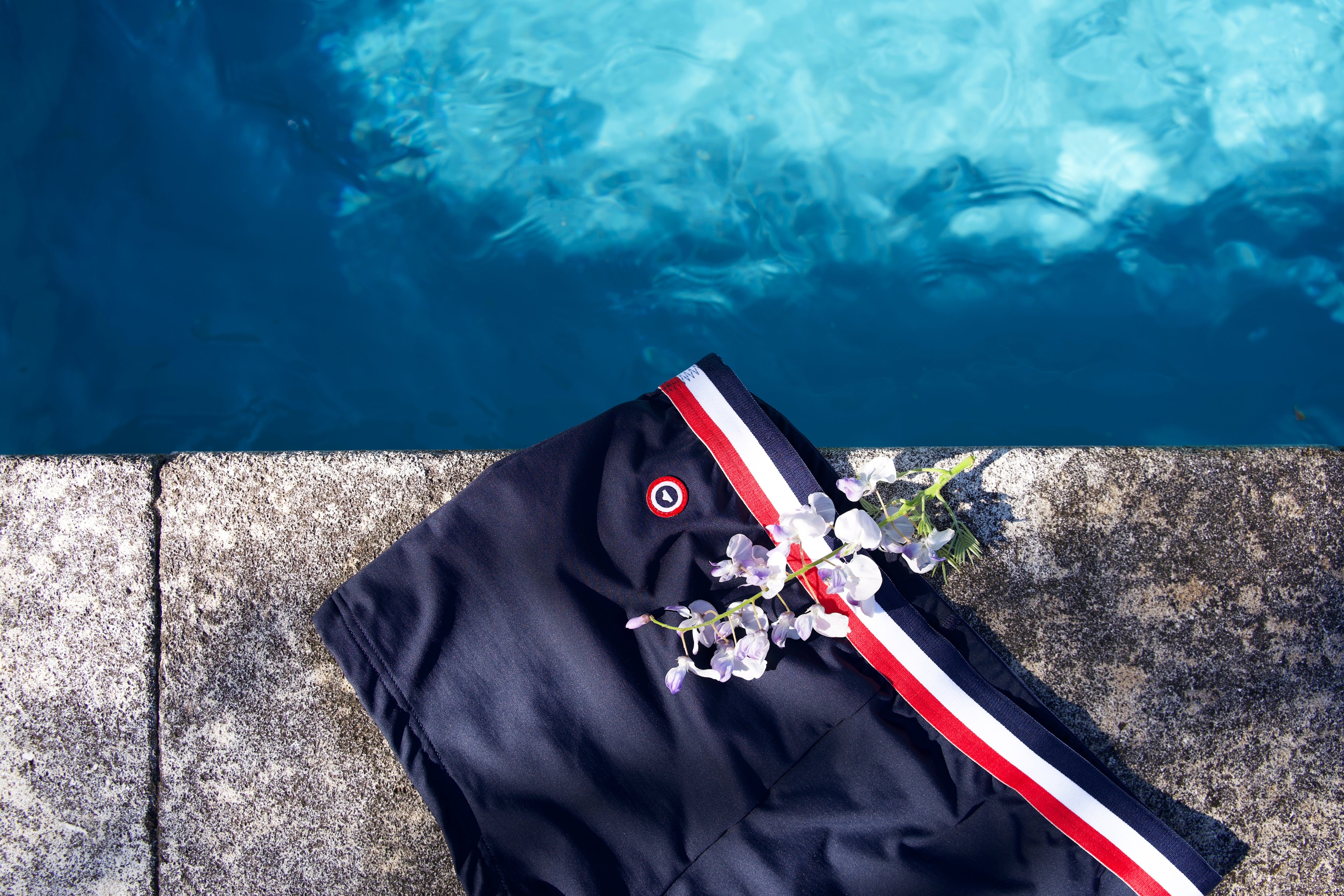 Gros plan d'un maillot de bain bleu marine à ceinture élastique tricolore orné d'une branche de fleurs blanches au bord de l'eau turquoise.