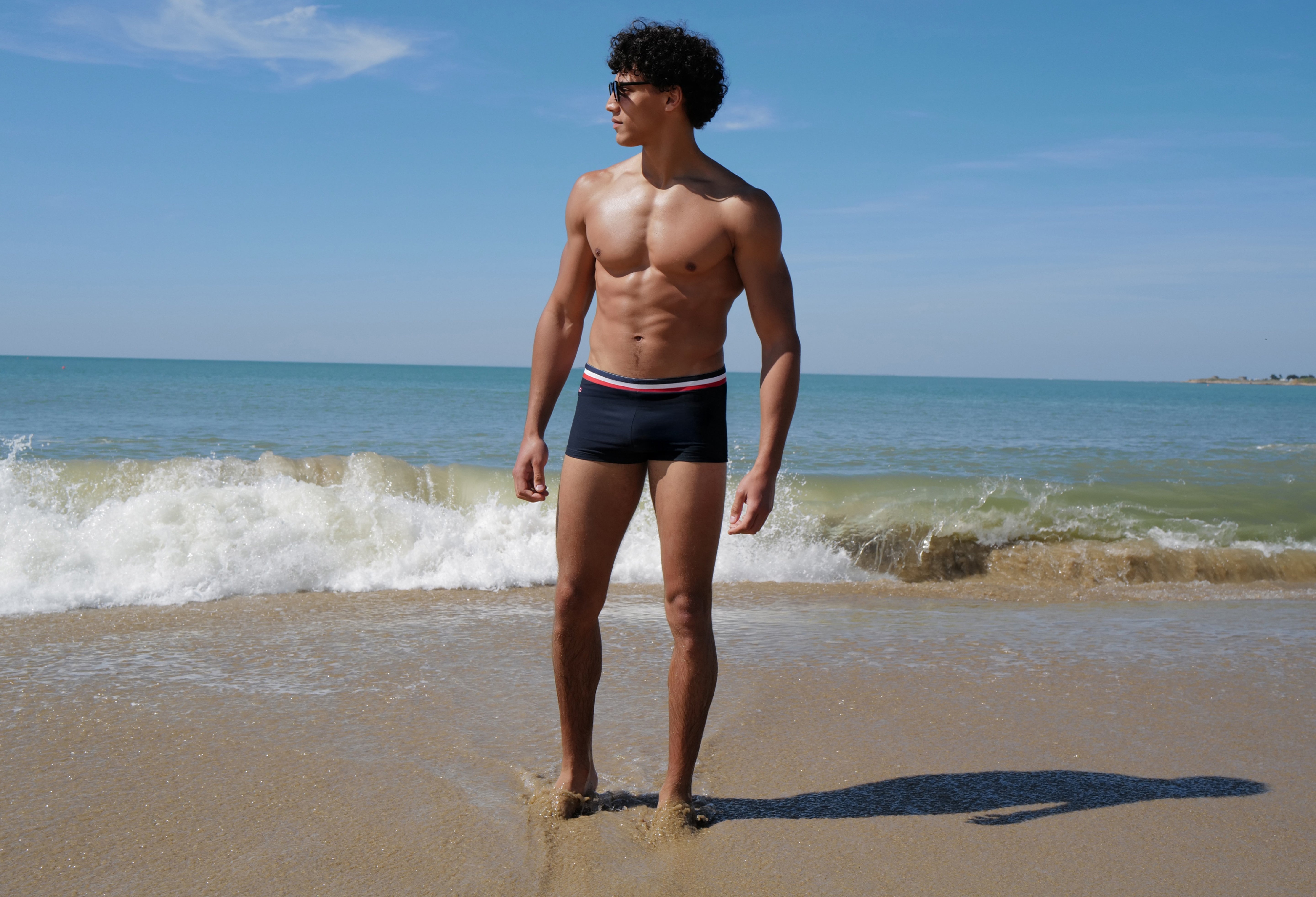 Homme athlétique portant un maillot de bain de type boxer de bain bleu marine avec liseré tricolore, debout sur une plage de sable face à l'océan.
