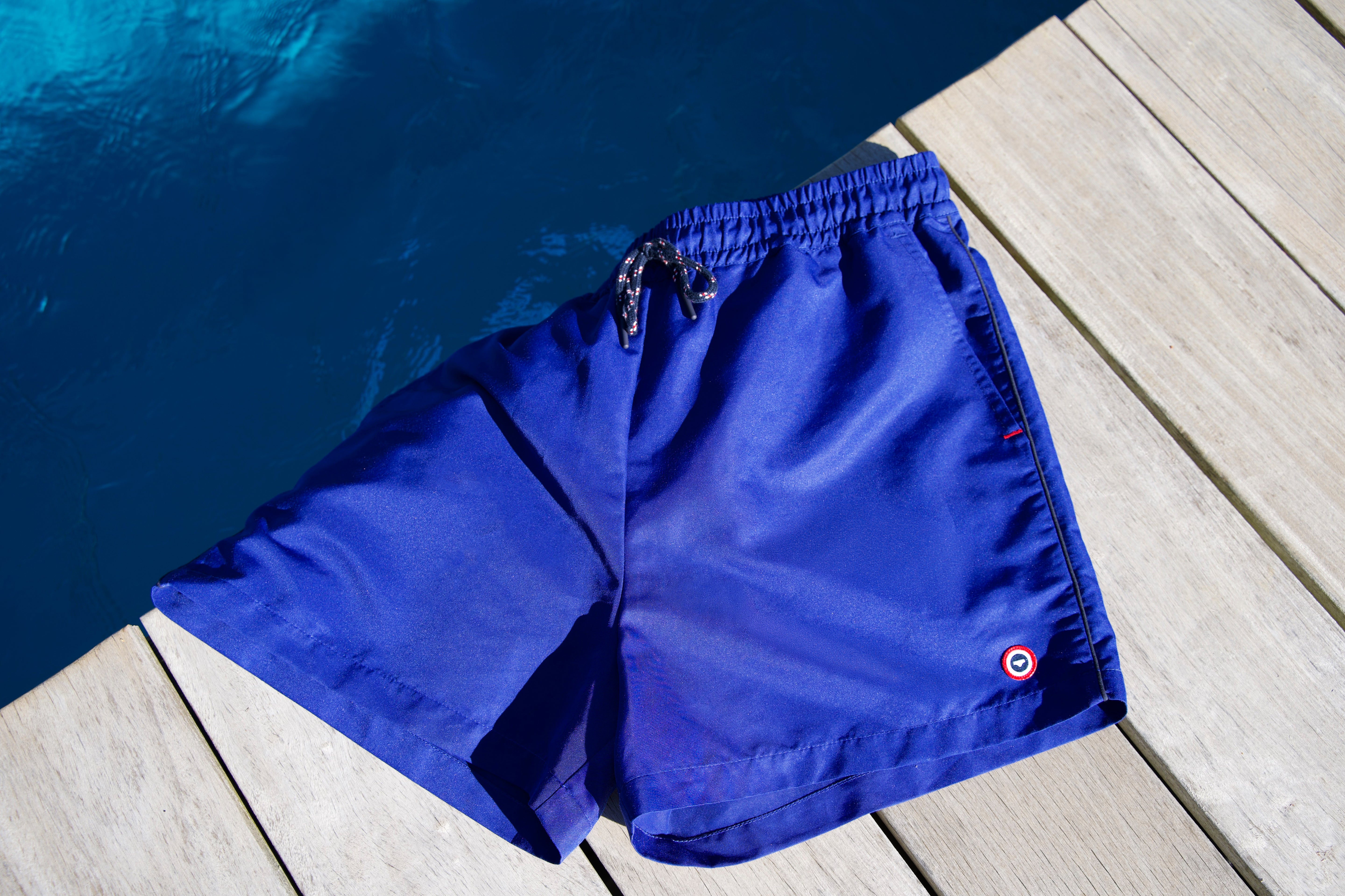 Short de bain bleu indigo pour homme avec cordon de serrage et logo, disposé sur un sol de piscine en bois clair.