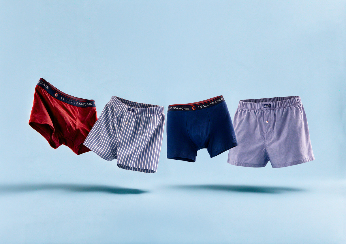 Quatre modèles de boxers pour hommes de la marque Le Slip Français flottant en l'air sur un fond bleu clair. De gauche à droite : un boxer rouge, un boxer à rayures bleues et blanches, un boxer bleu marine et un boxer lavande. Les ceintures des boxers rouges et bleu marine arborent fièrement l'inscription 'LE SLIP FRANÇAIS'.